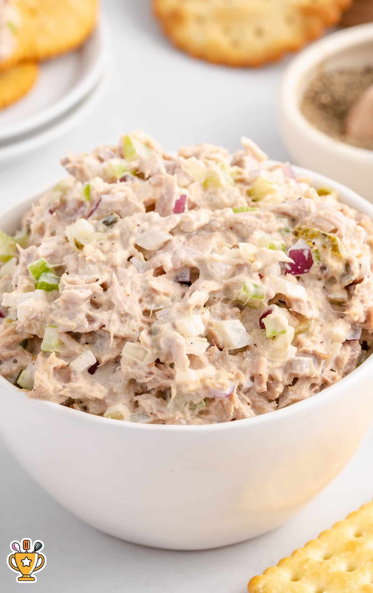 White bowl filled with homemade tuna salad, packed with chopped onions and celery, placed next to crackers and a bowl of pepper.