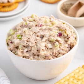 White bowl filled with creamy tuna salad surrounded by crackers, pepper, and more ingredients, ready for serving.