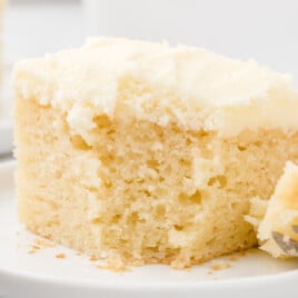A square slice of vanilla snack cake with white frosting served on a white plate with a fork beside it