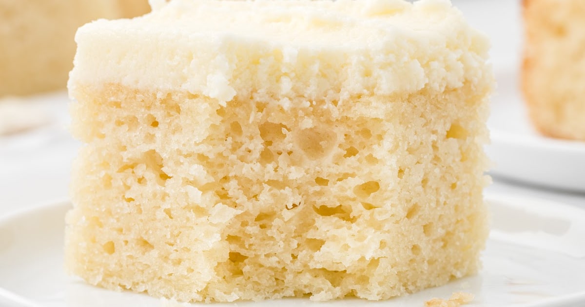 A partially eaten slice of vanilla snack cake with fluffy frosting , placed on a white plate.