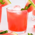 A refreshing watermelon cocktail served in a clear glass filled with ice, garnished with a wedge of watermelon and a sprig of fresh mint. The vibrant pink drink sits on a white surface, surrounded by extra watermelon slices and mint leaves.