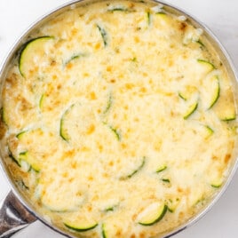 Cheesy zucchini gratin baked in a stainless steel skillet, topped with golden melted cheese and zucchini slices, on a white countertop.