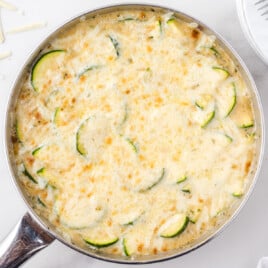 Creamy zucchini gratin baked in a stainless steel skillet, topped with melted golden-brown cheese and sliced zucchini rounds.