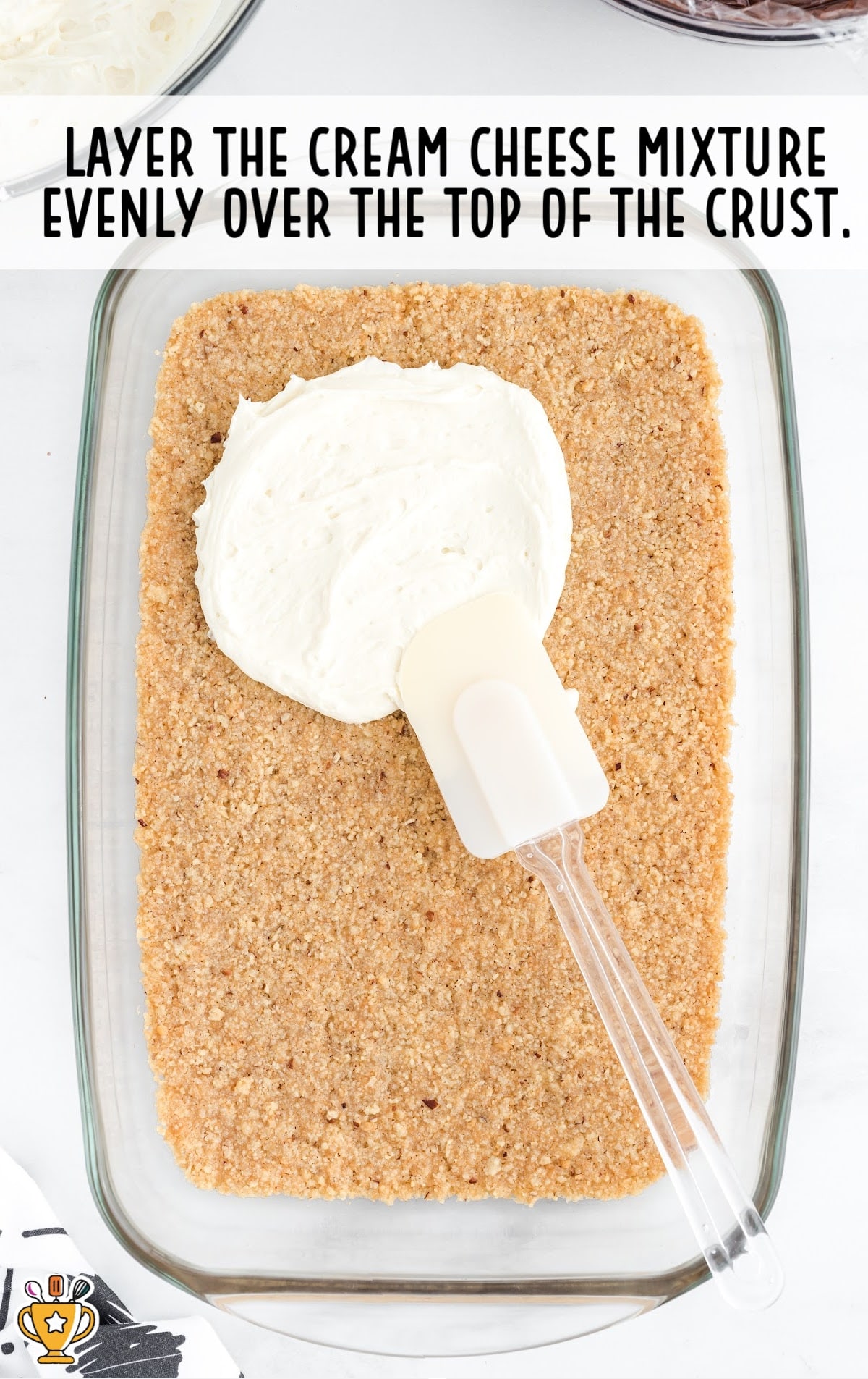 Photo of a spatula spreading a layer of fluffy cream cheese mixture over a baked pecan crust in a glass dish.