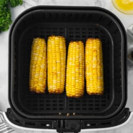 Four golden corn on the cob pieces cooked in an air fryer basket, with parsley, melted butter, and salt and pepper nearby.