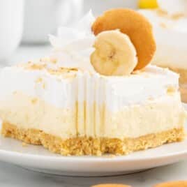 Close-up of a banana cheesecake bar with a graham cracker crust, creamy filling, whipped cream, banana slice, and Nilla wafer.