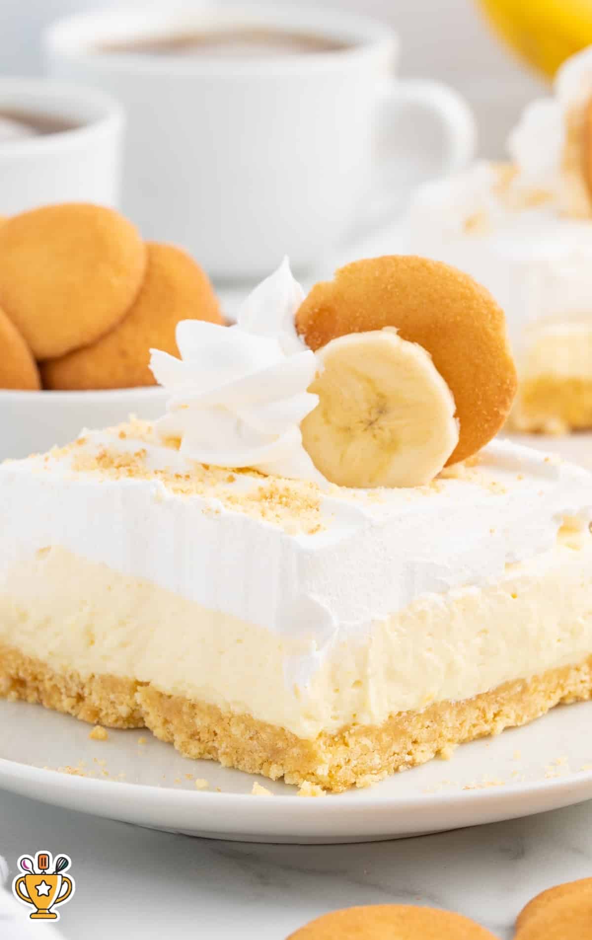 Banana cheesecake bar slice on a plate with creamy layers, whipped topping, banana slice, and Nilla wafer.
