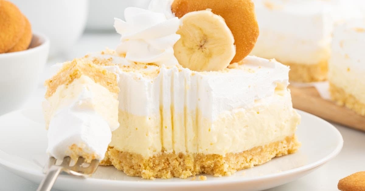 Banana cheesecake bar slice on a white plate with a fork holding a bite of creamy cheesecake and whipped topping.