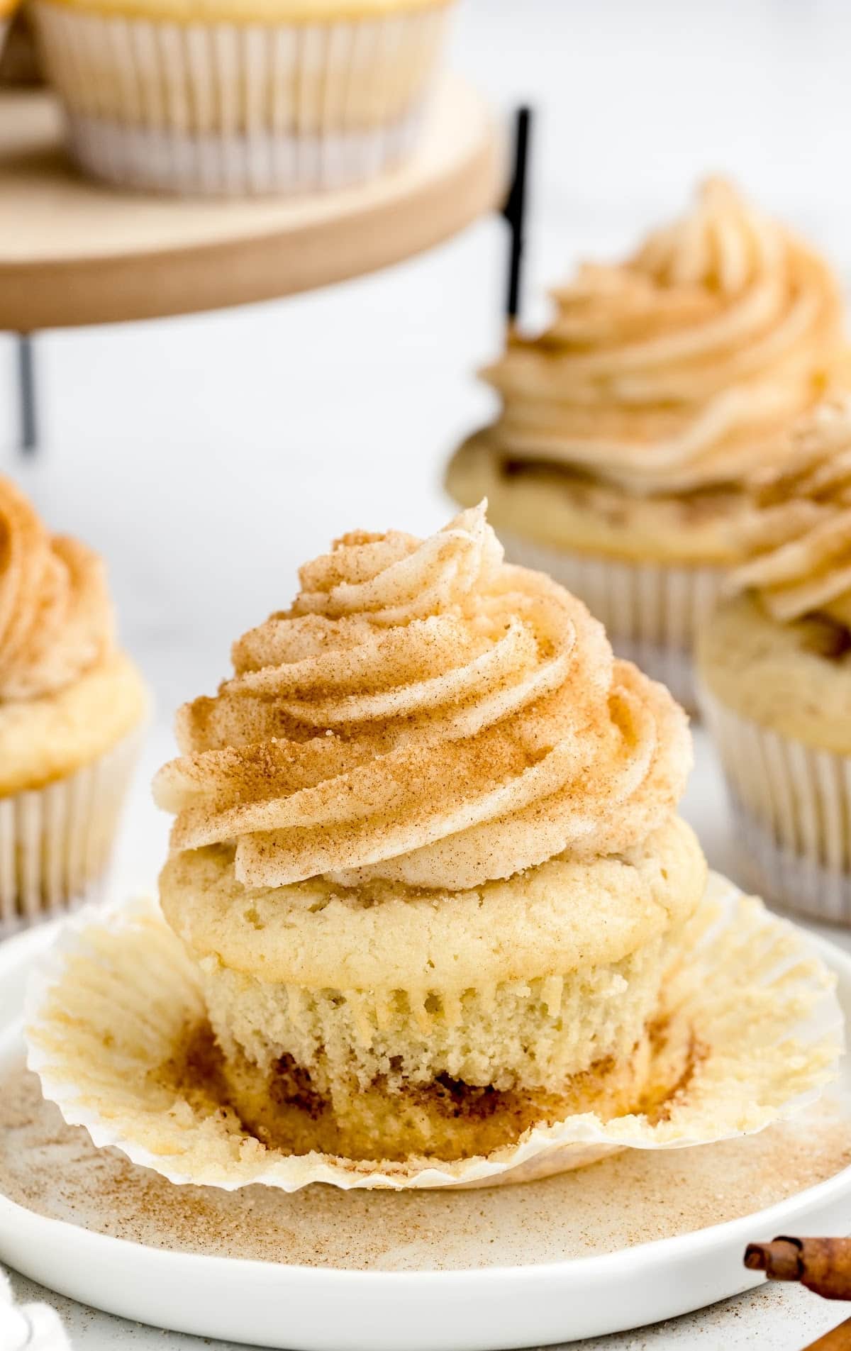 Cinnamon cupcake unwrapped on a white plate, with fluffy interior and a swirl of cinnamon sugar visible at the base. The cupcake is topped with a large swirl of frosting dusted with cinnamon, with more cupcakes displayed on a raised wooden stand in the background.