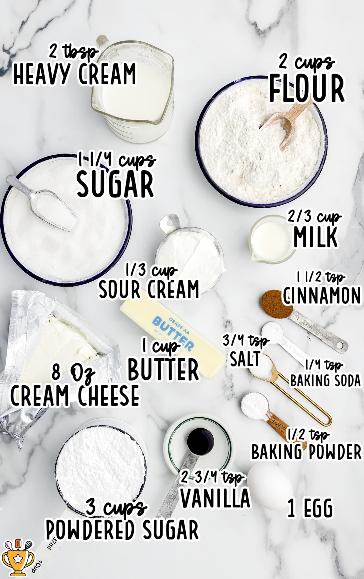 Flat lay of baking ingredients for cinnamon cupcakes, each labeled with text. Items include flour, sugar, butter, cream cheese, powdered sugar, sour cream, milk, heavy cream, vanilla, egg, cinnamon, salt, baking soda, and baking powder.