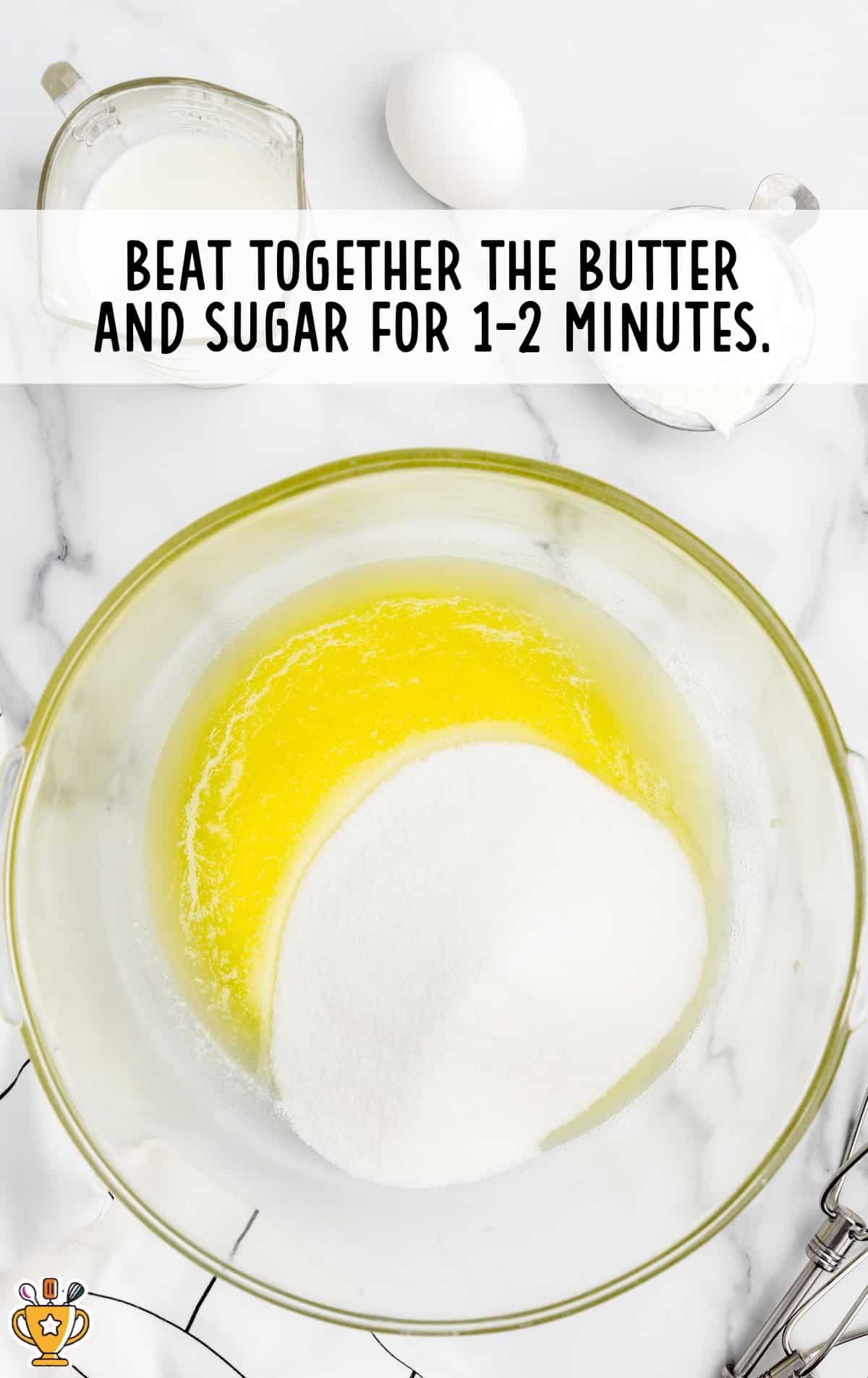 Overhead shot of a clear mixing bowl containing melted butter and granulated sugar, ready to be beaten together. Surrounding the bowl are a measuring cup of milk, a whole egg, and another cup of ingredients. Text overlay reads “Beat together the butter and sugar for 1–2 minutes.”