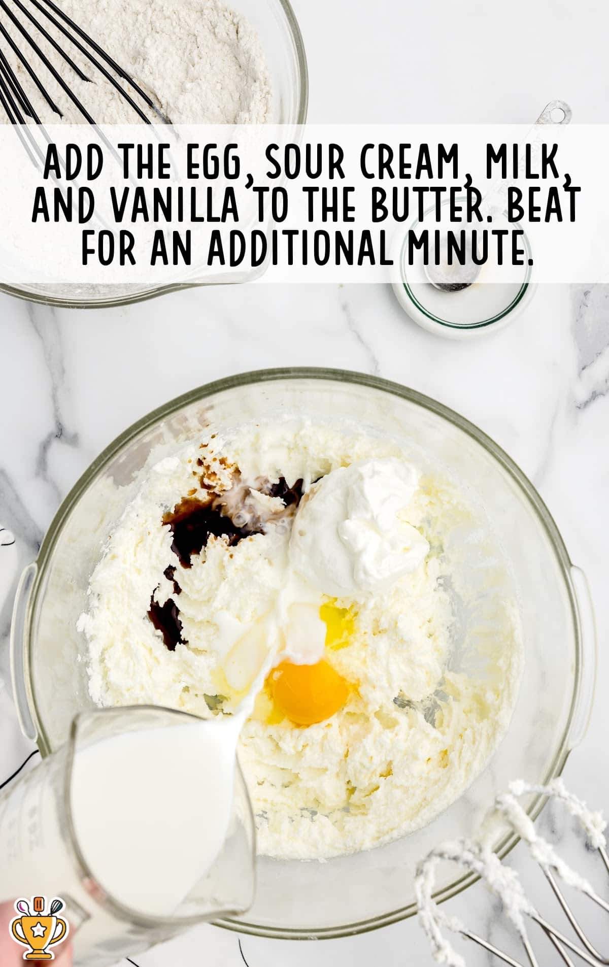 A large mixing bowl filled with a creamy mixture of butter and sugar, now combined with a raw egg, sour cream, vanilla extract, and milk. A hand is pouring milk into the bowl. Text overlay reads “Add the egg, sour cream, milk, and vanilla to the butter. Beat for an additional minute.”
