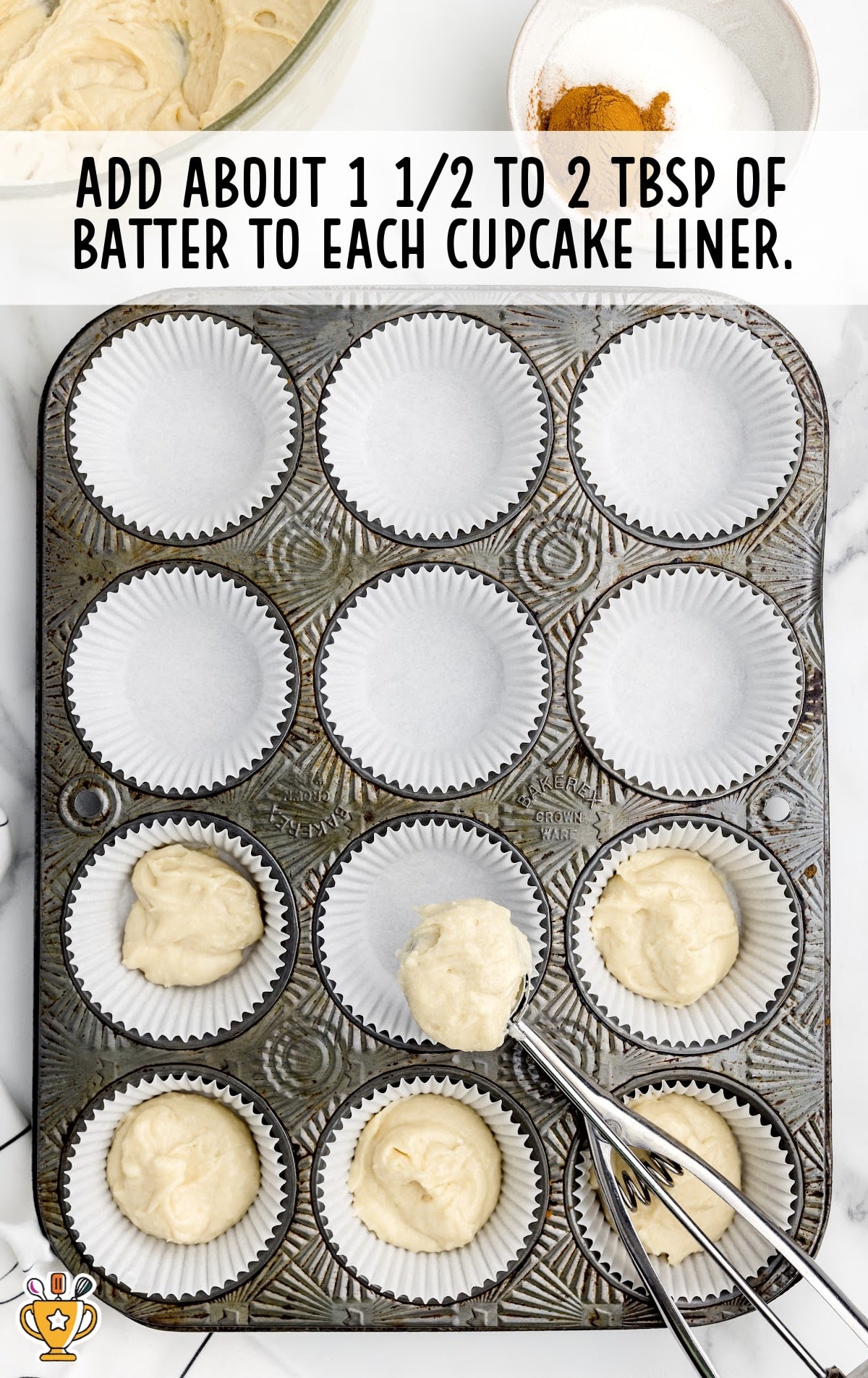 A muffin pan lined with white cupcake papers, some filled with spoonfuls of thick cupcake batter. A metal scoop is placing batter into the liners. Text overlay instructs, “Add about 1 ½ to 2 tbsp of batter to each cupcake liner.”