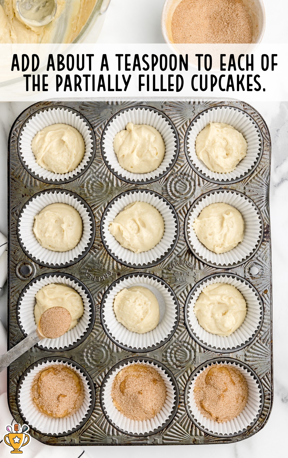 Muffin tin with cupcake liners partially filled with batter. A spoon is sprinkling a teaspoon of cinnamon sugar mixture into each cup. Several liners already have a coating of cinnamon sugar. Text overlay reads “Add about a teaspoon to each of the partially filled cupcakes.”
