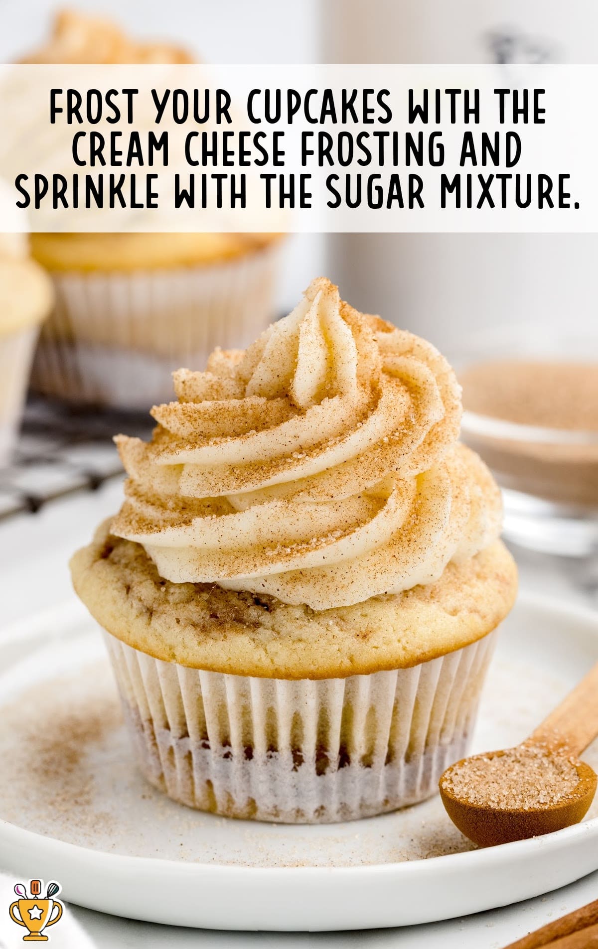 Finished cinnamon cupcake topped with a swirl of cream cheese frosting and dusted with cinnamon sugar. The cupcake sits on a white plate beside a wooden spoon filled with extra cinnamon sugar. Text overlay reads “Frost your cupcakes with the cream cheese frosting and sprinkle with the sugar mixture.”