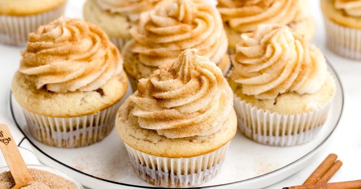 Plate full of cinnamon cupcakes with tall swirls of frosting generously sprinkled with cinnamon sugar. A small wooden spoon with extra cinnamon sugar sits in the foreground, alongside two cinnamon sticks.