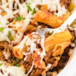 Overhead view of baked ziti in a white crockpot, topped with melted mozzarella and chopped herbs, with a serving spoon inside.