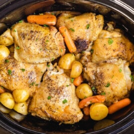Top-down view of cooked chicken thighs in a slow cooker with baby potatoes and carrots, garnished with fresh parsley.