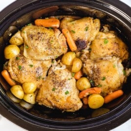 Slow cooker filled with golden brown chicken thighs, baby carrots, and potatoes, perfectly cooked and garnished with fresh parsley.