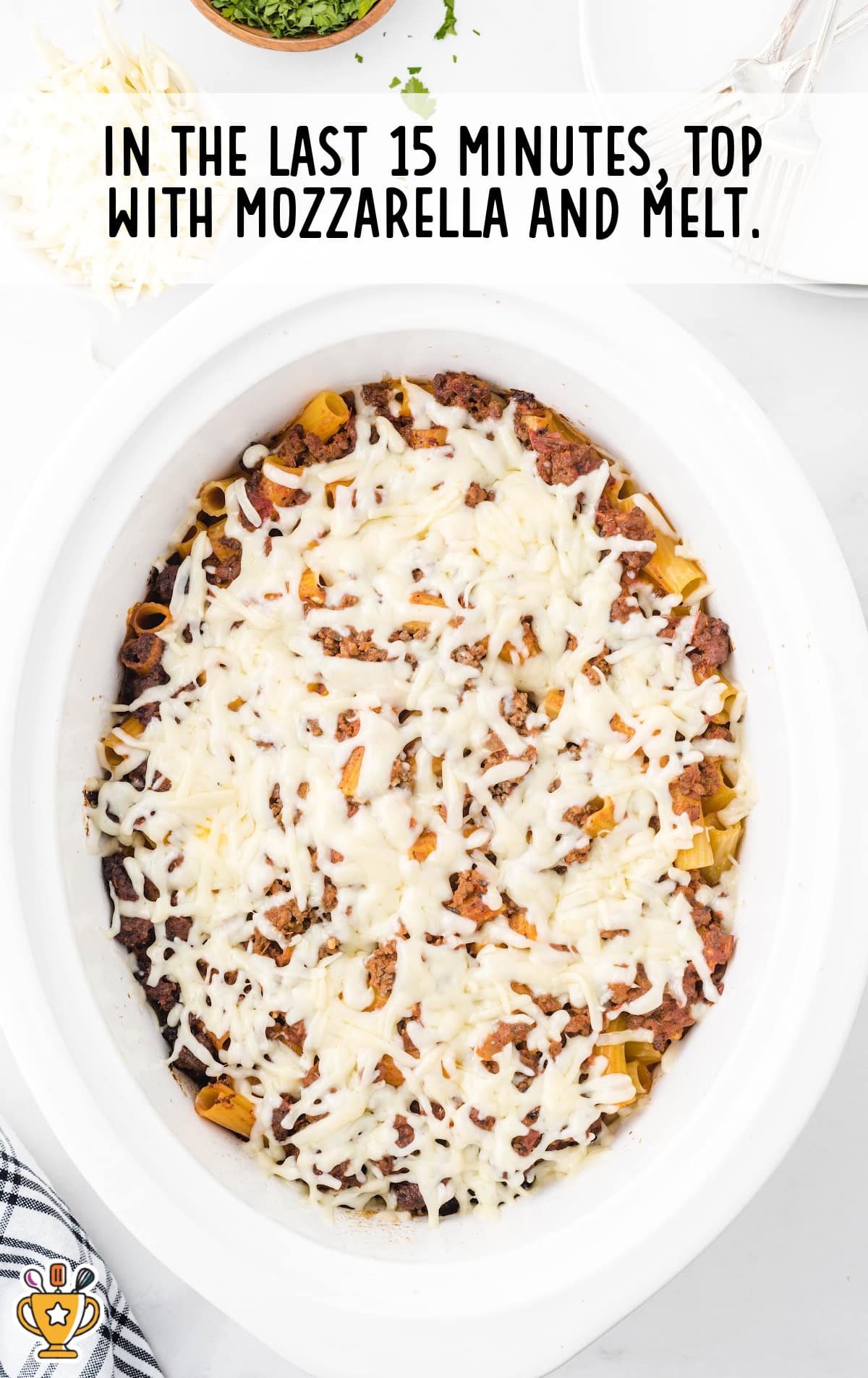 Crockpot filled with baked ziti topped with melted mozzarella cheese, ready to serve, with forks and herbs nearby.