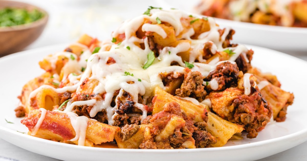 Split view of a plated serving and crockpot full of cheesy baked ziti with a metal spoon inside, garnished with parsley.