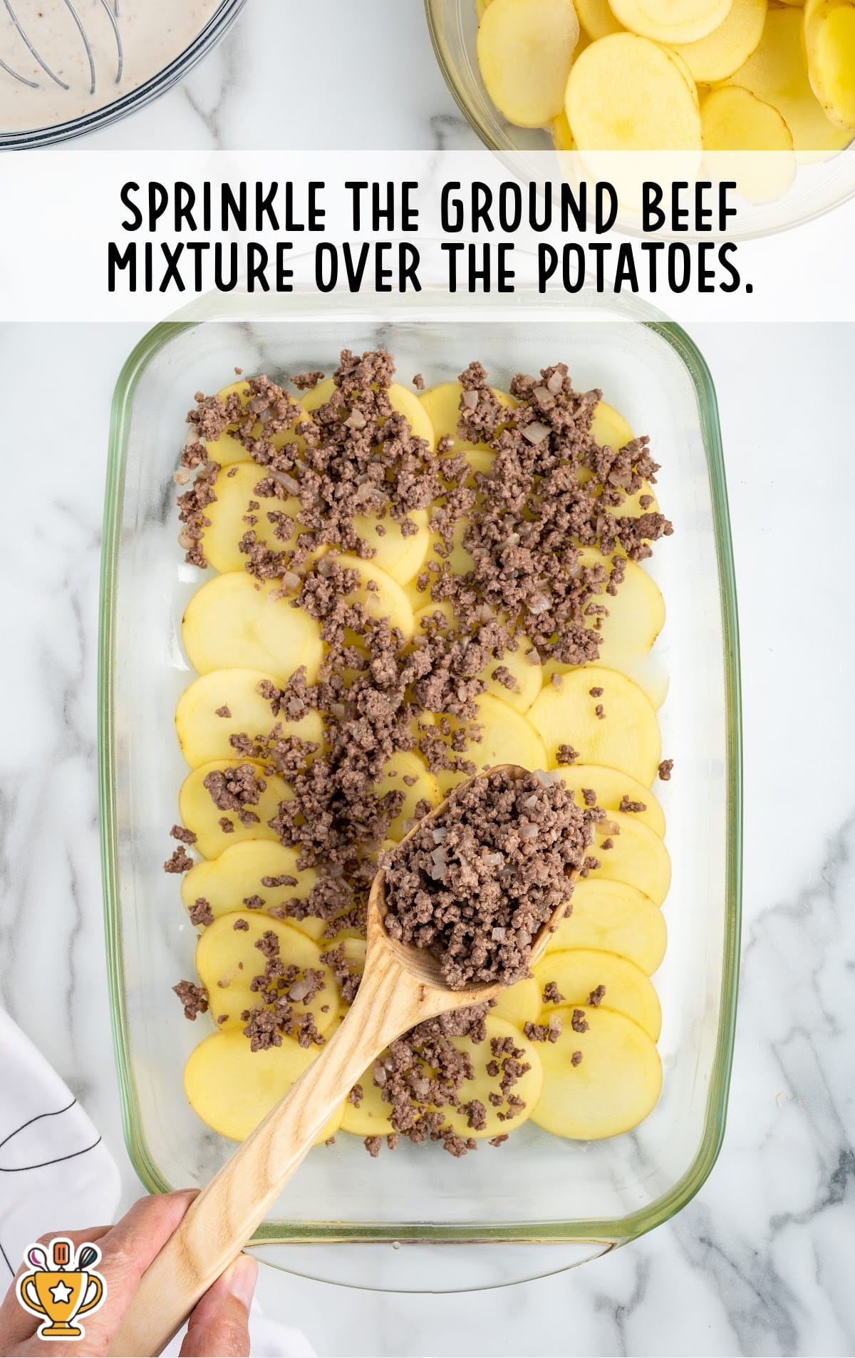 Sliced potatoes arranged in a glass baking dish with cooked ground beef mixture being sprinkled on top with a wooden spoon.