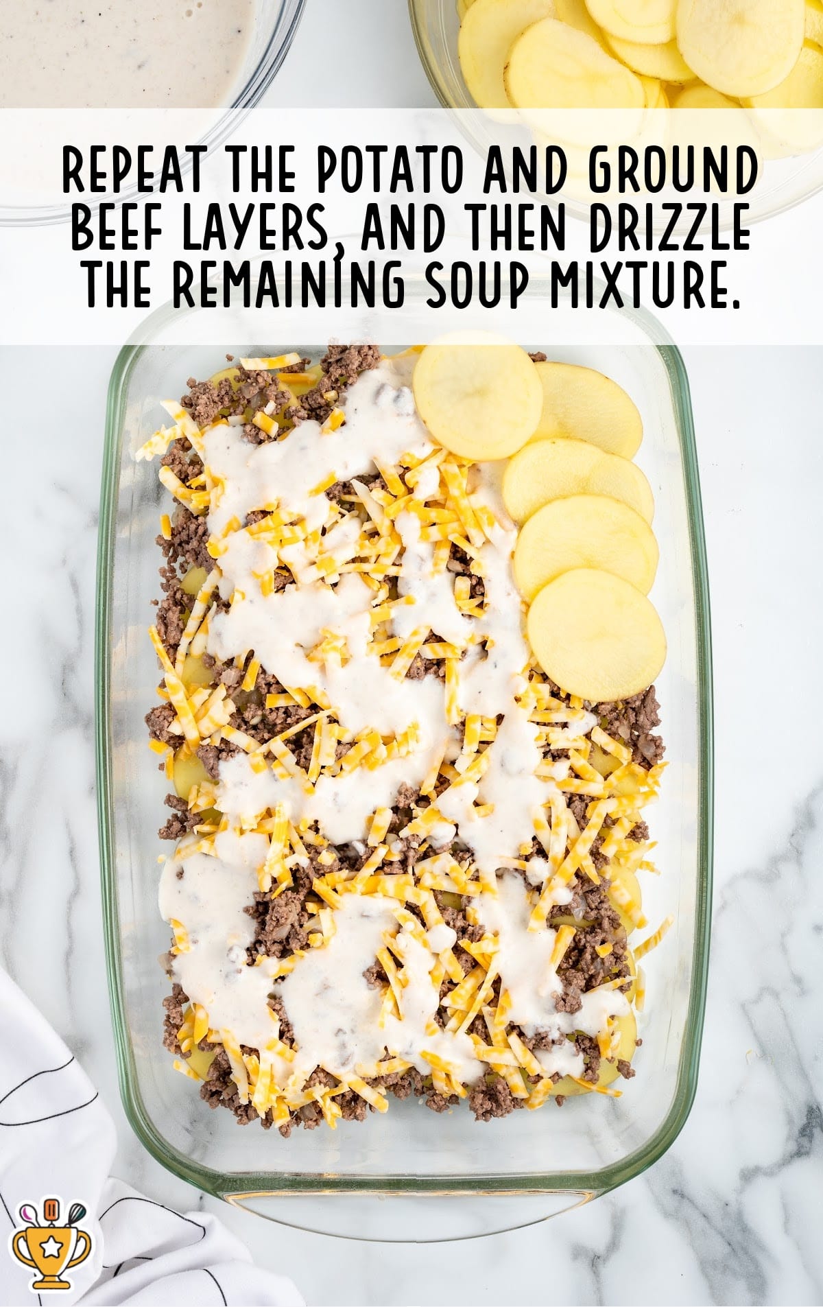 Glass baking dish layered with potatoes, ground beef, shredded cheese, and creamy soup mixture ready for baking.