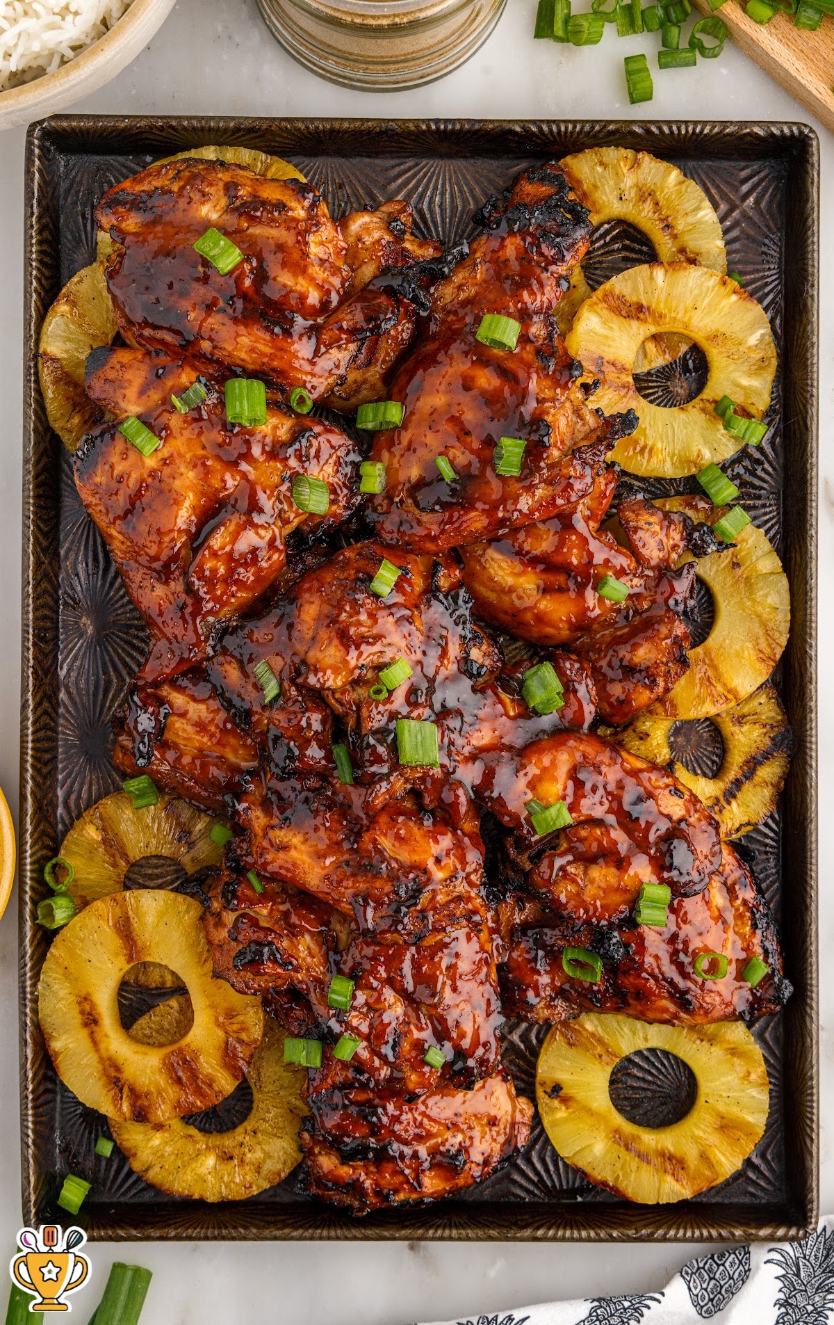 Overhead view of grilled huli huli chicken glazed with a glossy pineapple soy marinade, topped with chopped green onions, and served on a tray with golden grilled pineapple rings.