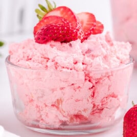 Glass bowl filled with creamy pink strawberry Jello salad topped with fresh strawberry slices, surrounded by whole strawberries on a white surface.