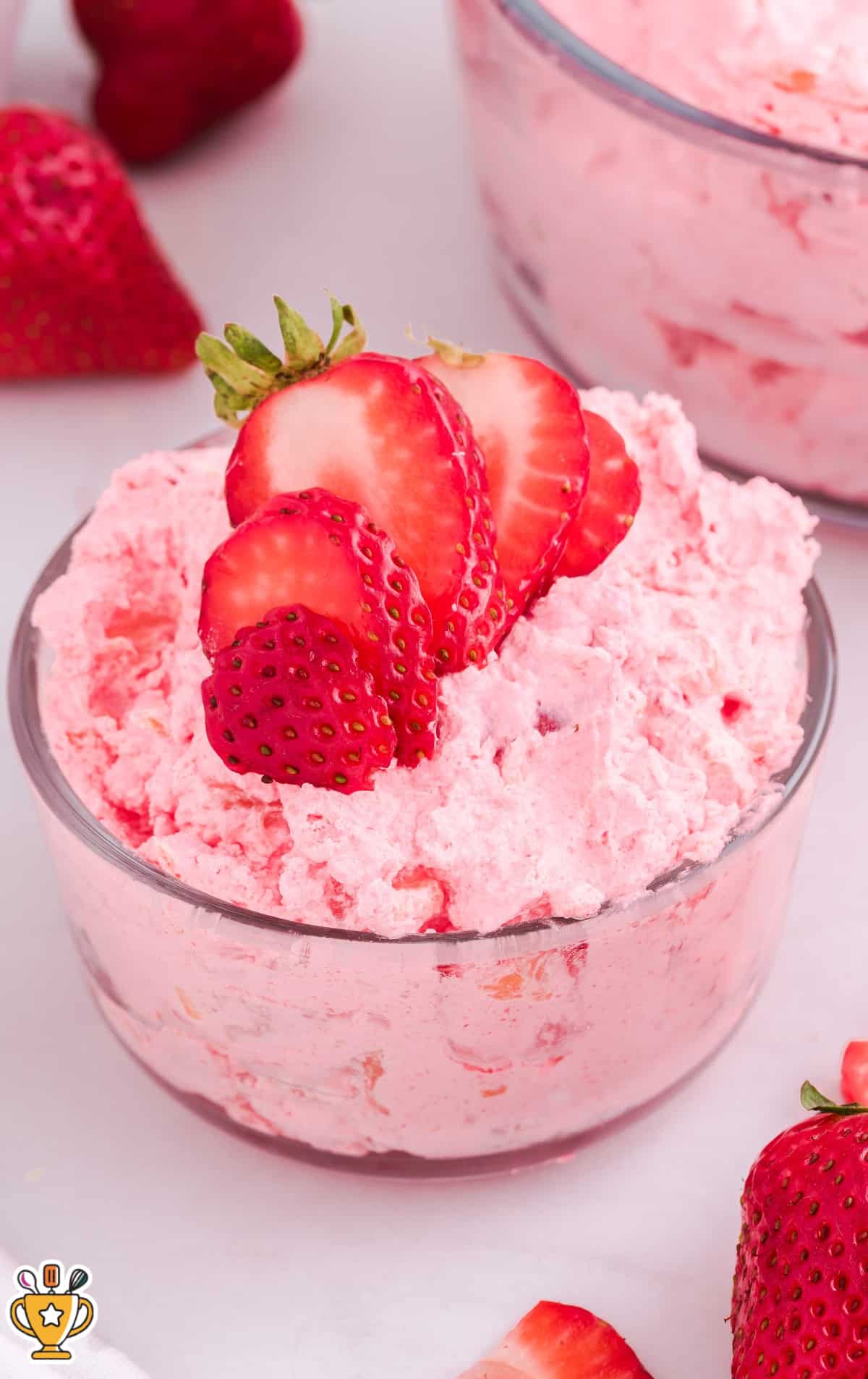 Glass serving bowl filled with strawberry Jello salad topped with three fanned strawberry slices, set against a soft background with scattered strawberries.