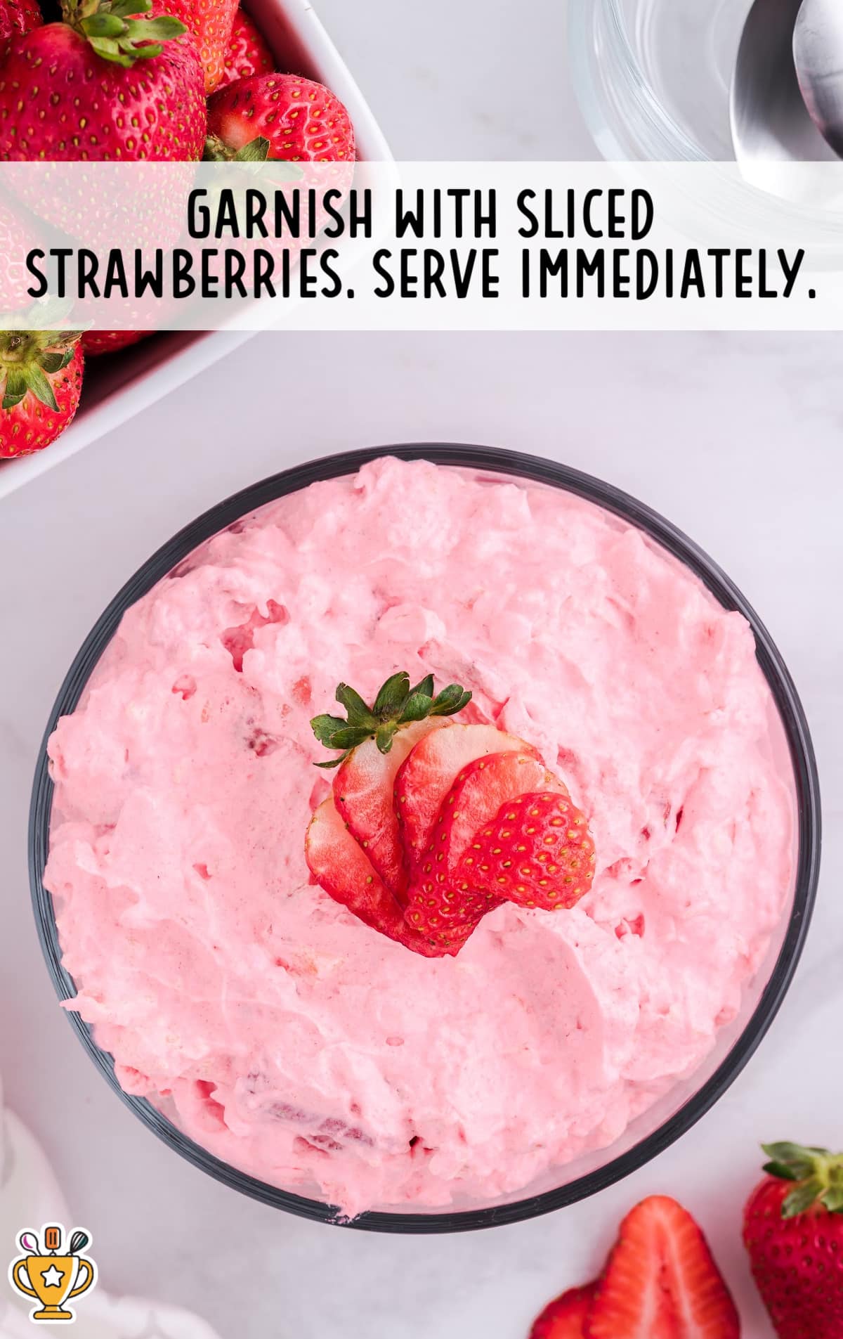 Top-down view of a glass bowl filled with fluffy pink strawberry Jello salad, garnished with fresh strawberry slices. Surrounded by whole and halved strawberries, with the caption &ldquo;Garnish with sliced strawberries. Serve immediately.&rdquo;