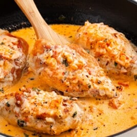 Golden chicken breasts cooking in creamy tomato Parmesan sauce in a skillet, with a wooden spoon lifting one piece of chicken.