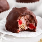 A close-up of a Martha Washington candy with a bite taken out, showing a creamy pink coconut filling with chopped pecans and maraschino cherries, coated in a smooth layer of chocolate and resting in a white candy paper liner.