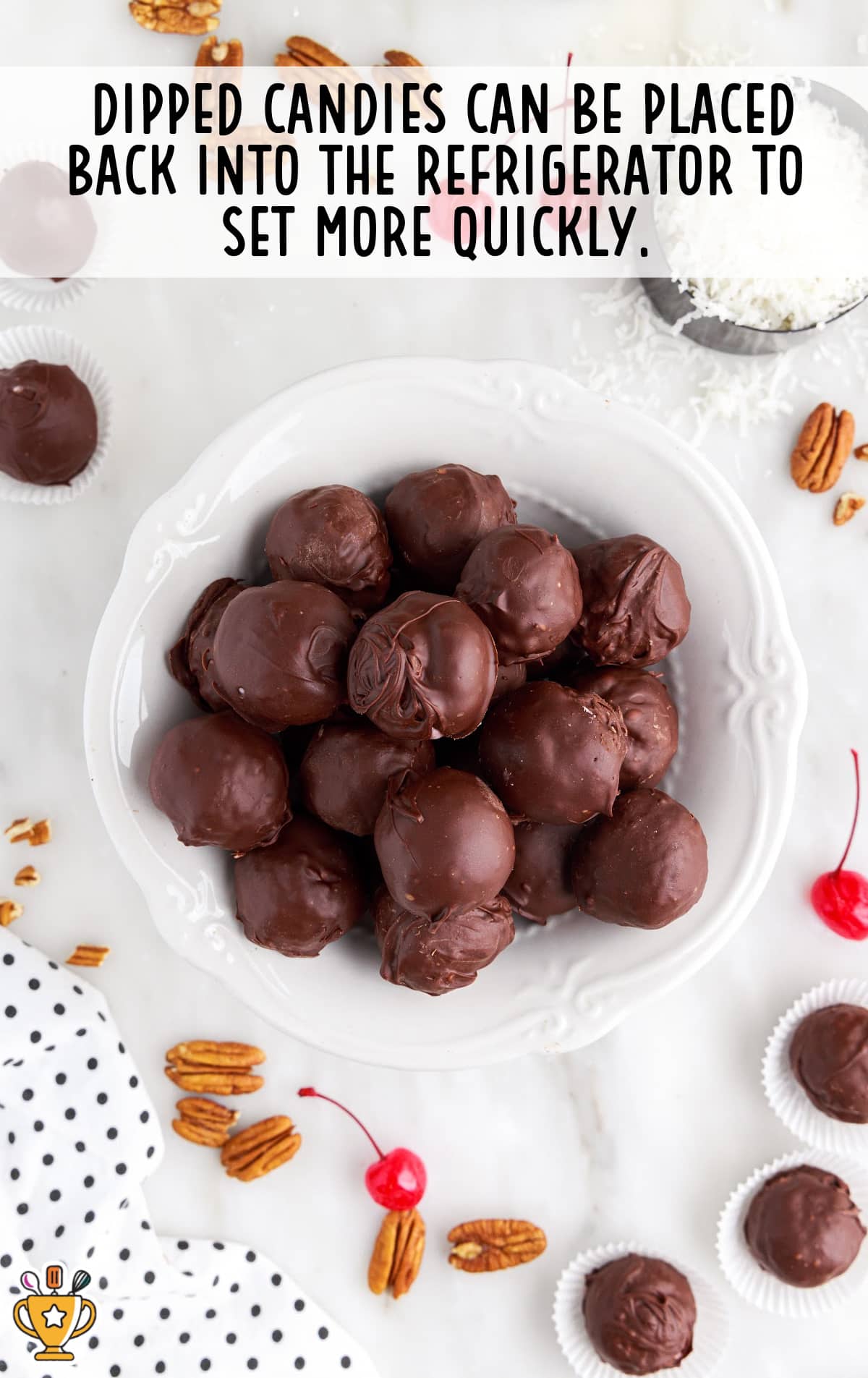 Finished Martha Washington candies piled in a white bowl, surrounded by scattered pecans, cherries, and candy cups on a white surface.