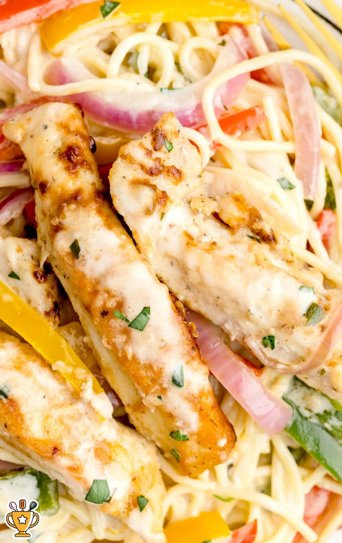 Top-down close-up of golden chicken strips over creamy spaghetti noodles with sautéed onions and peppers, garnished with herbs.