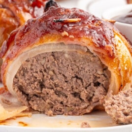 Close-up of a cut-open onion bomb on a white plate. The inside reveals a juicy meat filling, with a dipping bowl of barbecue sauce just behind.