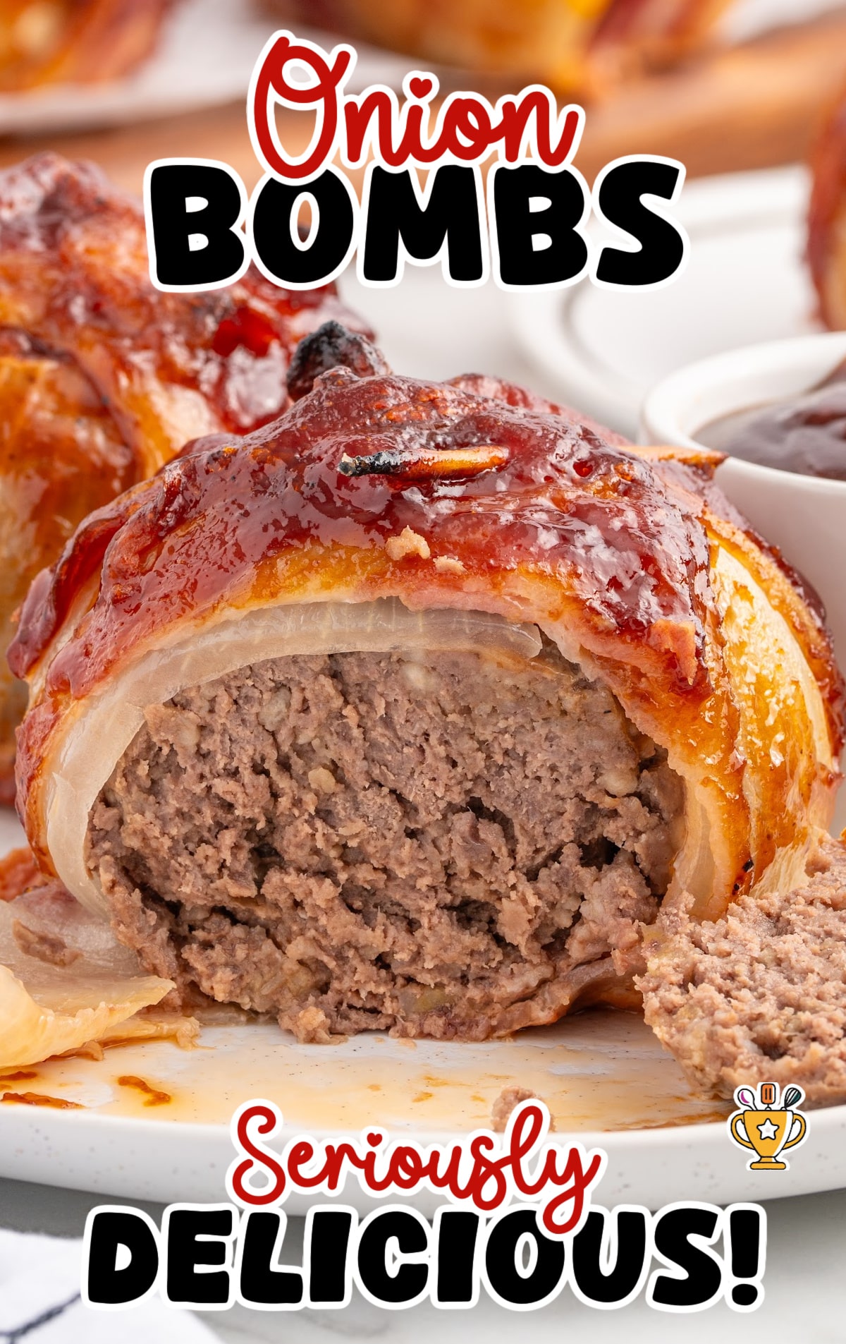 Close-up of a bacon-wrapped onion bomb cut in half, showing the moist ground beef center. Barbecue sauce glistens on top, and a dipping bowl is seen behind it.