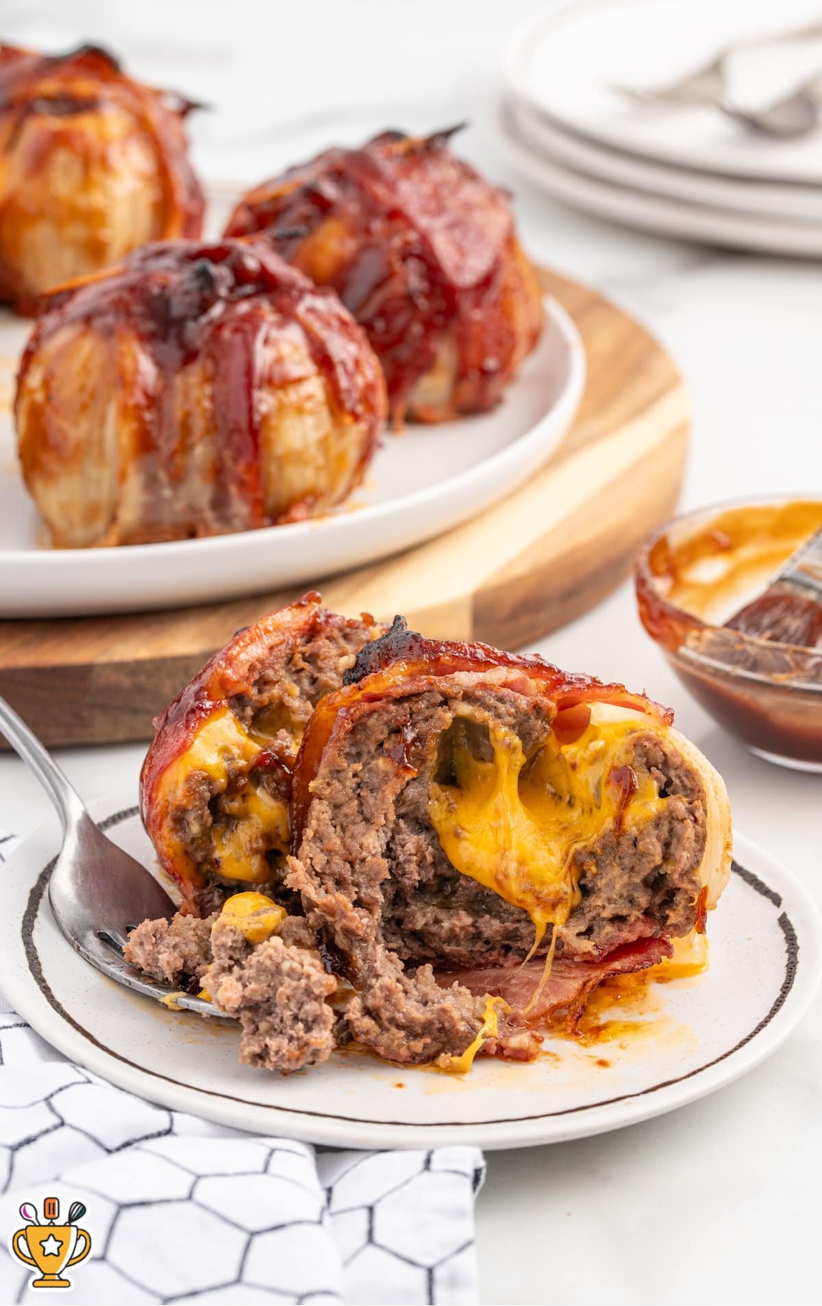 Cheddar cheese melts from the center of a sliced open onion bomb on a white plate. A fork rests nearby and more wrapped bombs are in the background, ready to serve.