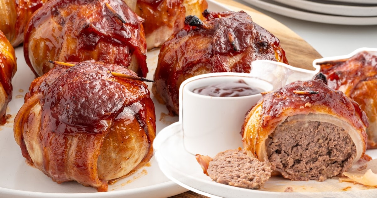 A platter of glazed, bacon-wrapped onion bombs with one cut open beside a bowl of barbecue sauce for dipping.