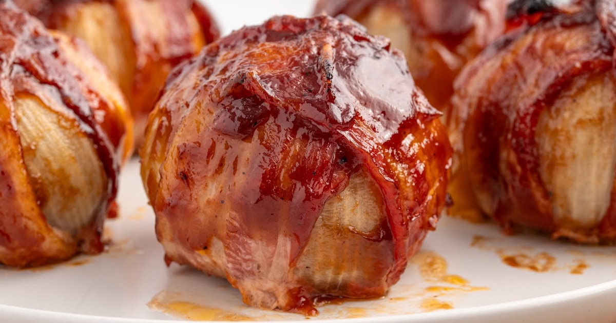 Close-up of golden-brown, bacon-wrapped onion bombs resting on a white plate, coated with sticky barbecue glaze.
