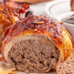 A juicy onion bomb has been sliced open to reveal a tender meat filling. The crispy bacon is glazed in a sticky barbecue sauce, and a small dish of sauce sits in the background.