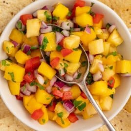 White bowl filled with pineapple mango salsa topped with a silver spoon, surrounded by crunchy tortilla chips on a serving tray.