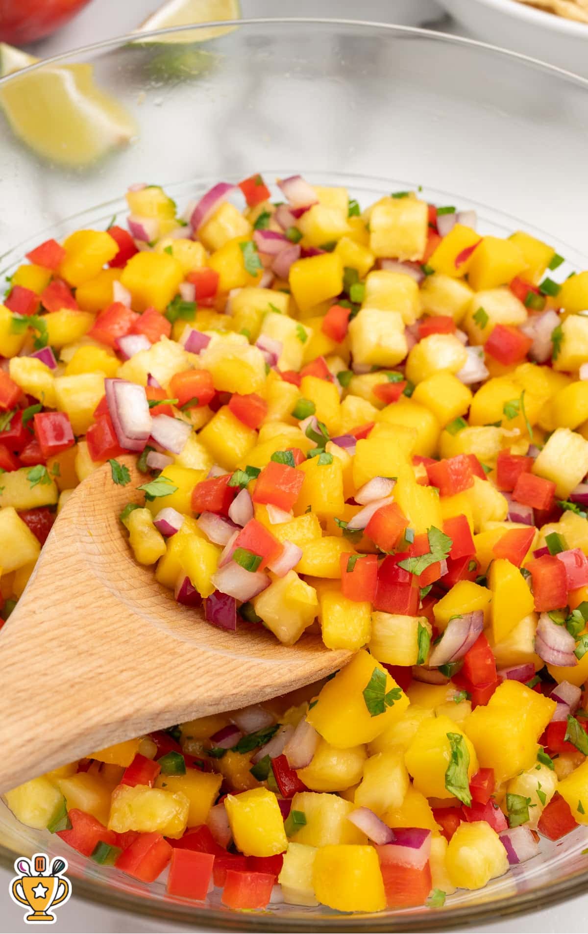 A wooden spoon mixing a colorful blend of chopped pineapple, mango, red bell pepper, red onion, jalapeño, and cilantro in a glass bowl.