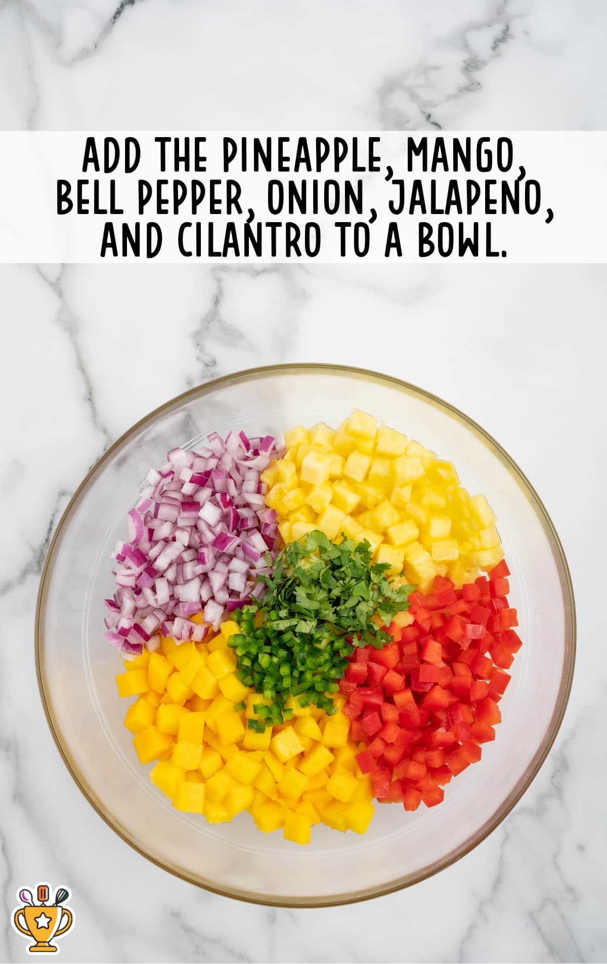 A clear glass mixing bowl filled with separated piles of diced pineapple, mango, red onion, bell pepper, jalapeño, and cilantro on a white marble background.