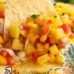 A tortilla chip scooping pineapple mango salsa from a hollowed pineapple, filled with vibrant yellow, red, and green ingredients.