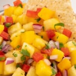 Bright pineapple mango salsa in a white bowl with a tortilla chip dipped in, surrounded by festive text perfect for summer parties.