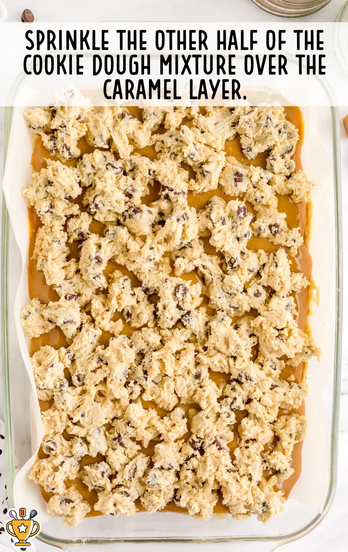 Glass baking dish with caramel layer topped with chunks of cookie dough mixture, ready for baking.