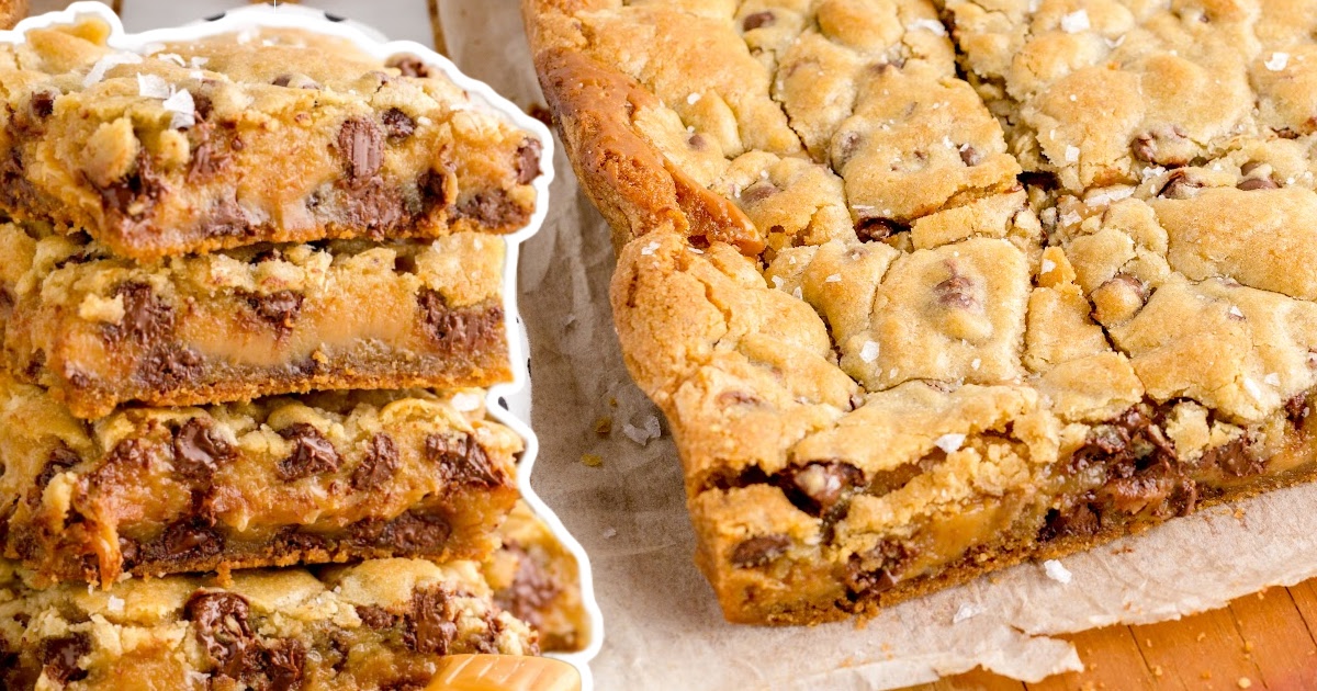 A full baking dish of salted caramel chocolate chip cookie bars next to a tall stack of cut squares showing gooey caramel layers.