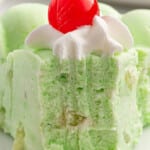 a close-up of a creamy green slice of seafoam salad topped with a cherry, highlighting lime gelatin as an ingredient.