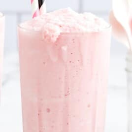 Two tall glasses of creamy pink strawberry frosty with striped straws, one glass in focus with froth overflowing slightly over the rim.
